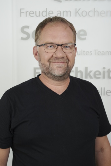 Mitarbeiterbild stehender Mann mit Brille in schwarzem T-Shirt, der in die Kamera lächelt