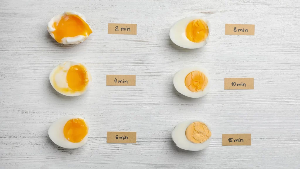 Weißer Holzhintergrund auf dem 6 halbierte, gekochte Eier liegen mit unterschiedlich hartem Eigelb. Neben den Eiern stehen die Kochzeiten auf kleinen braunen Zetteln.