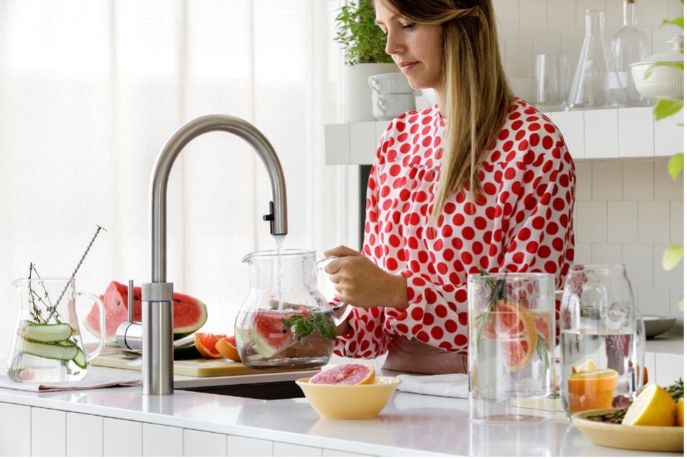 Stehende junge Frau an einer Küchenspühle füllt sich frisches Wasser aus der Armatur in eine Glaskaraffe
